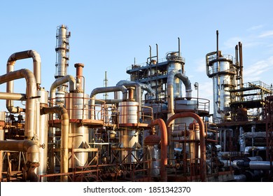 Old Distillation Column Towers And Reactors Under Blue Evening Sky Background At Chemical Plant. Exterior Of Silver Metal Rusty Enterprise With Copyspace.