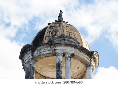 An old dome structure with weathered columns and birds perched atop against a partly cloudy blue sky