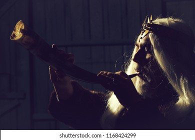 Old Druid Bearded Man With Long Beard On Serious Face And Hair In Fur Coat And Crown With Gem Stones Jewellery On Wooden Background Holding Animal Antler Or Horn.