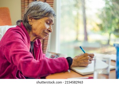 Old Elderly Asian Indian Woman Sitting Writing In A Journal Or Notebook, UK. Depicts Memory Loss And Keeping A List Or Diary.