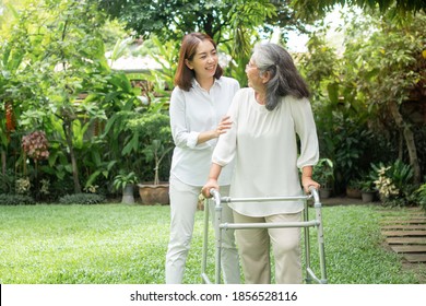 An Old Elderly Asian Woman Uses A Walker And Walking In The Backyard With Her Daughter.  Concept Of Happy Retirement With Care From A Caregiver And Savings And Senior Health Insurance