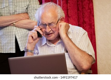 Old Elderly Senior Man On Phone At Laptop Computer At Risk To Cyber Attack And Online Bank Fraud