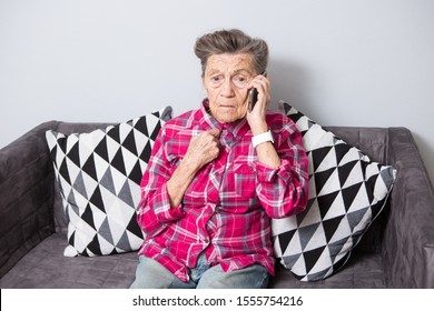An old elderly woman grandmother with gray hair sits at home on the couch using the hand phone, a telephone conversation to hear the bad news. Emotion fear scare.