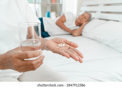 Old Elderly Woman Holding Medicine And Water On Hand On The Bed In The Bedroom And Her Husband Sleeps Beside. Concept Of Sleep Problems In The Elderly And Use Of Sedatives And Sleep Medicine Addiction