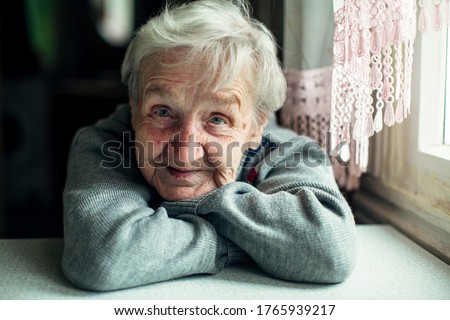 An old elderly woman, smiling portrait, looking at the camera, sitting in the kitchen.