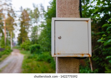 Old Electric Shield On A Concrete Pole In The Countryside Near A Dirt Road. The Old Electric Shield, A Rusty Metal Box Hangs On The Pole Electric Pole Of The Garden