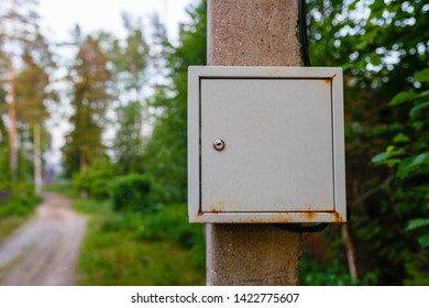 Old Electric Shield On A Concrete Pole In The Countryside Near A Dirt Road. The Old Electric Shield, A Rusty Metal Box Hangs On The Pole Electric Pole Of The Garden