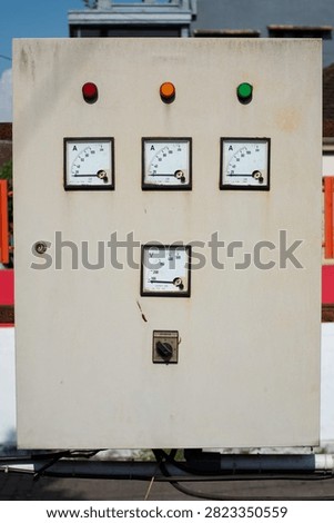 Old electrical control panel box with analog gauges, meters, switches, and colorful indicator lights on a wall. Vintage industrial power system or generator control dashboard.