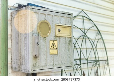 Old electrical meter box with high voltage warning sign on a house wall. Weathered metal electricity distribution box mounted outdoors. Industrial utility cabinet with a yellow lightning bolt danger symbol, showing signs of aging, dust, and rust on a residential building exterior.