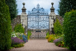 An Old And Elegant Wrought Iron Gate Stands At The End Of An Entranceway Lined With Hedges, Trees And Flowers