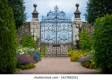 An old and elegant wrought iron gate stands at the end of an entranceway lined with hedges, trees and flowers