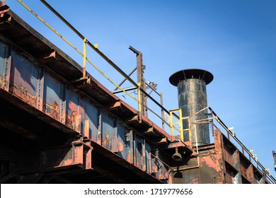 Old Elevated Train Tracks Heavy With Rust And Peeling Paint, Smoke Stack Behind As Part Of An Abandoned Industrial Plant, Blue Sky, Horizontal Aspect