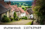 Old English limestone houses with thatched roofs with green fields countryside in the background. Gold Hill houses on a cloudy day behind flowers in Shaftesbury, Dorset, UK. Photo with selective focus