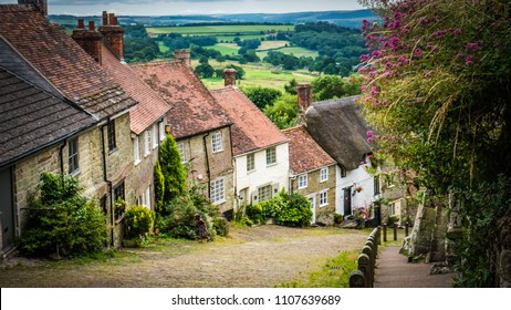 Old English limestone houses with thatched roofs with green fields countryside in the background. Gold Hill houses on a cloudy day behind flowers in Shaftesbury, Dorset, UK. Photo with selective focus - Shutterstock ID 1107639689