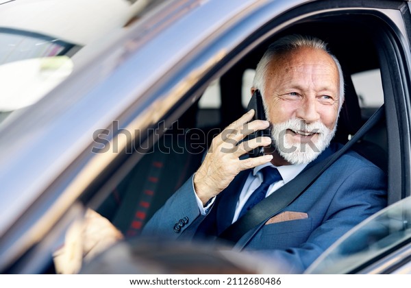 An old\
executive has a phone call while driving a car. A happy senior\
businessman sits in his car with his hand on the steering wheel and\
has a phone call while looking through the\
window.