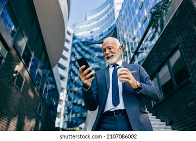 An old executive reading message on the phone at the business center. Low angle view of a happy senior businessman standing at the business center and reading a message on the phone. - Shutterstock ID 2107086002