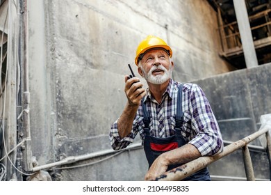 An old factory worker with a helmet on his head, leans on the railing inside of the factory and communicates with his coworkers over the walkie-talkie. Workers talking with walkie-talkie. - Powered by Shutterstock