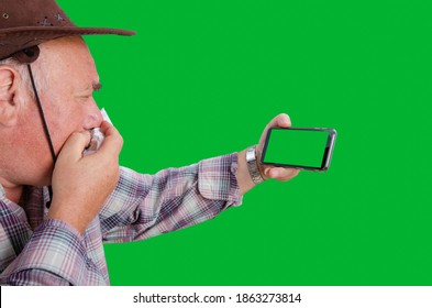 An Old Farmer Can't Help Coughing While Talking To Someone Via Video Chat On A Chroma Key Smartphone. It Looks Like The Old Man Has Contracted The Coronavirus. Telemedicine Topic. Green Background.