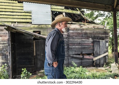 Old farmer standing in front of a rustic dilapidated wooden barn with his hands in his pocket staring thoughtfully off to the side