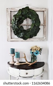 Old Farmhouse Window Decorated With A Homemade Magnolia Leaf Wreath Hung On An Interior Wall Over Rustic Half Moon Table With Candles And Flowers Decor.