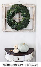 Old Farmhouse Window Decorated With A Homemade Magnolia Leaf Wreath Hung On An Interior Wall Over Rustic Half Moon Table With Heirloom Pumpkins.