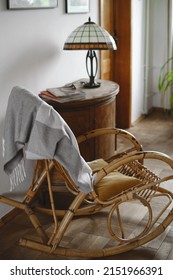 Old Fashion Wicker Rocking Chair With Blanket In Cozy House, Lamp On Wooden Cabinet. Place For Reading, No People, Home Plant Near Window. Comfortable Vintage Living Room Interior