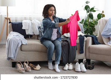 Old Fashioned Clothes. Portrait Of Thoughtful Black Woman Making Revision Of Her Clothing At Home, Sitting On Couch With T-Shirt In Hands, Free Space