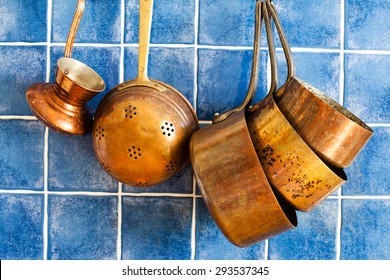 Old fashioned copper kitchenware, utensils. Hang on iron hooks against blue tile wall. Coffee maker, colander,  antique pans: casserole, stewpot, pot.