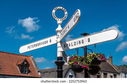 Old fashioned Holland County Council road sign in Crowland, Lincolnshire showing Peterborough, Spalding and Thorney. Also showing a thatched roof cottage, bright hanging baskets and blue sky.