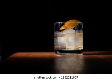 Old Fashioned With An Orange Peel Or It Can Become A Rob Roy Cocktail. Whiskey On The Rocks In A Clear Glass On Top Of A Wooden Table And Black Background 