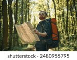 Old fashioned paper map in hands. Bearded man is in the forest at daytime.