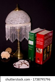 An Old Fashioned Table Lamp With Glass Shade, Two-non Fiction Books And Spectacles, With A Night-time Setting And Black Background.   