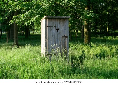 Old Fashioned Toilet Building With Wooden Door Standing Outside Among The Grass With Trees In The Background.