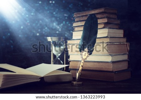 Old feather quill ink pen with inkwell and old books over wooden desk in front of black wall background. Conceptual photo on history, fantasy, education and literature topic