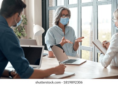 Old female mentor coach wearing face mask training young workers at group meeting in reopen office. Middle aged businesswoman teacher working with students at university class. Social distance concept