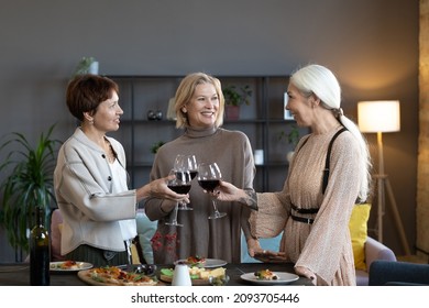 Old Friends Smiling And Toasting With Glasses Of Red Wine During Dinner Party In The Living Room At Home