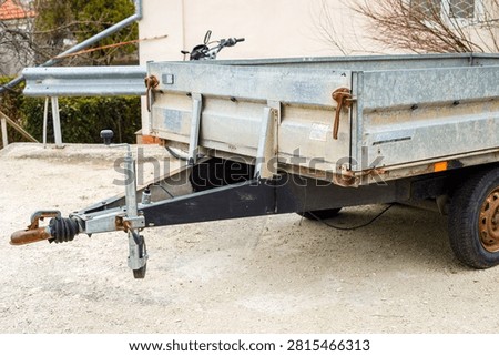 An old galvanized utility trailer sits parked on a gravel surface, the trailer's weathered metal sides showing rust and years of wear, with the trailer's tow hitch and wheel visible in the foreground and a residential building softly blurred behind the trailer in the background