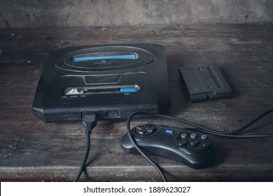 An Old Game Console In Black, In The Dust On A Wooden Table, Next To The Joystick And Cartridge.