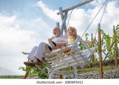 Old Grandfather And Young Child Grandson Swinging In Garden Outdoors. Grand Dad And Grandson Sitting On Swing In Park. Family Relationship Grandfather And Child.