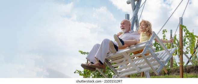 Old Grandfather And Young Child Grandson Swinging In Garden Outdoors. Grand Dad And Grandson Sitting On Swing In Park. Grandfather And Grandchild, Spring Banner.