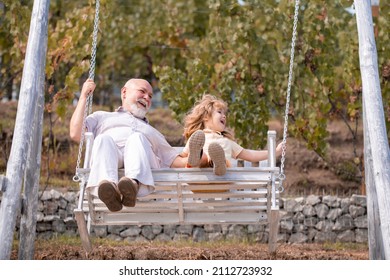 Old Grandfather And Young Child Grandson Swinging In Garden Outdoors. Grand Dad And Grandson Sitting On Swing In Park.