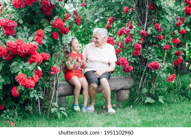 an old grandmother tells the girl funny stories from her childhood sitting on a bench made of logs in the garden under an arch of roses.