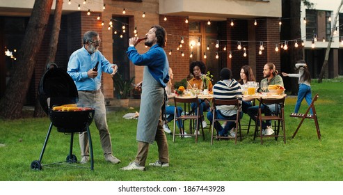 Old Gray-haired Father And Middle-aged Son Standing Outdoor At Barbecue On Back Yard, Drinking Beer And Talking. Caucasian Adult Man With Retired Senior Dad Having Nice Talk, Cooking Food On Fresh Air