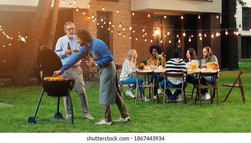 Old gray-haired father and middle-aged son standing outdoor at barbecue on back yard, drinking beer and talking. Caucasian adult man with retired senior dad having nice talk, cooking food on fresh air