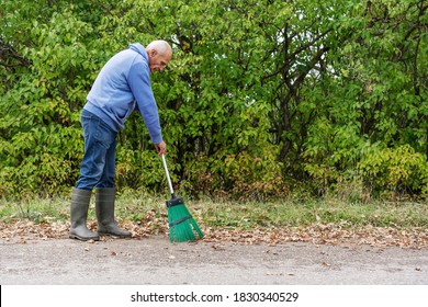 Old Grey Haired Gardener Man Sweeping Dry Foliage On Asphalt Road Side View