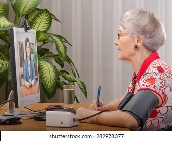 Old Grey Haired Woman Is Measuring Blood Pressure By Herself During Virtual Doctor Visit. Glasses Senior Woman Sitting Opposite Monitor. On The Screen, Telehealth Doc Is Consulting Her. Side Shot