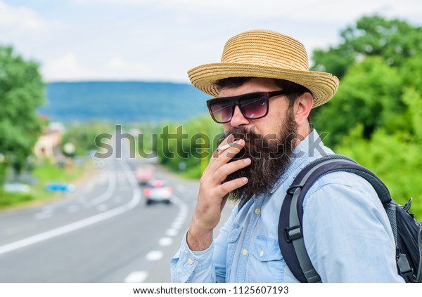 Old\
habit. Man with beard and mustache in straw hat smoking cigarette,\
road background defocused. Traveler stylish hipster take brake with\
cigarette. Smoking cigarette before long\
journey.