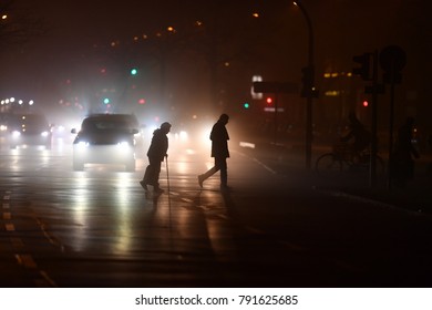 An Old And Handicapped Woman Crossing A Street In Berlin At Night. A Bright And Shiny Street Scene In Berlin-Germany. The Silhouettes Of Walking People.