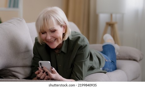 Old Happy Caucasian Grandmother 60 Years Senior Woman 50s Lady Blonde Elderly Female Lies On Couch At Home Enjoys Remote Mobile Conversation Video Call Conference Chat Smiling Talking To Phone Camera