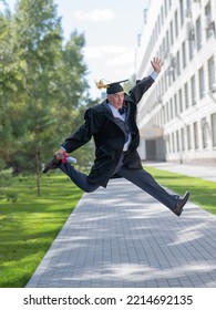 Old Happy Man In Graduation Gown Jumping Outdoors And Holding Diploma. Vertical.